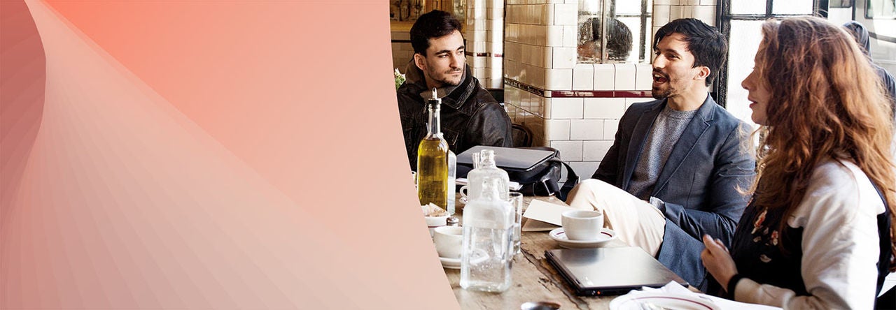 Three people are sitting at a wooden table in a cafe, engaged in conversation. The table has various items including a laptop, notebooks, coffee cups, and glass bottles.