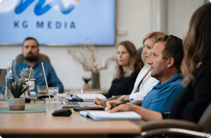 Photograph of a KG Media business meeting with five people seated around a conference table, focusing on a man in a blue shirt speaking. A large screen in the background displays "KG MEDIA," and the table holds laptops, notebooks, and plants, indicating a professional discussion or presentation.