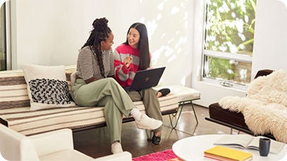 Two people sitting on a couch indoors. One person is holding a laptop and talking to the other.