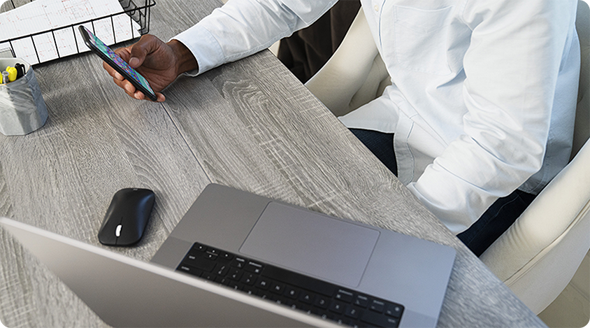 Person sitting at a desk holding a smartphone near an open laptop and work materials.