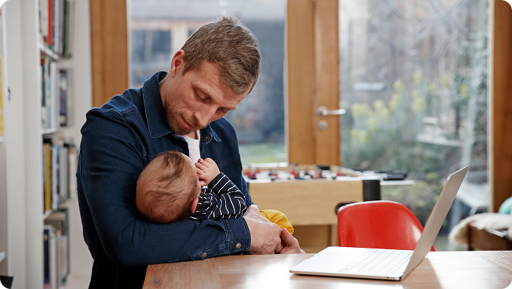 Man holding baby and shopping online on a laptop at home, highlighting anytime shopping.