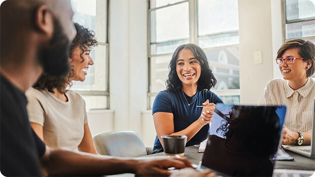 Four people sitting at a desk, smiling and using laptops.