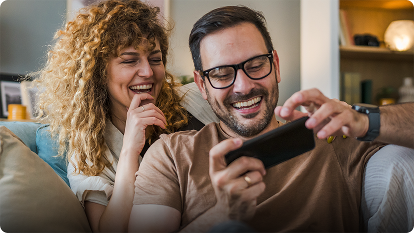 Two people sitting on a couch, one holding a smartphone horizontally while the other leans in closely, with shelves and home decor visible in the background.
