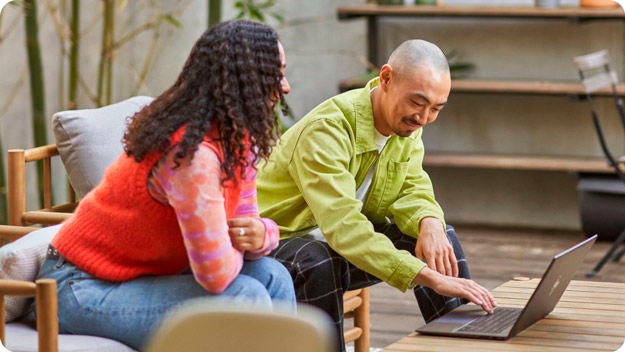 Two people seated at a wooden table, one using a laptop. Indoor plants and modern furniture suggest a casual work environment.