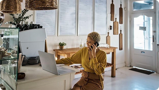A person with short, curly hair sits at the counter of a well-lit café, engaged in a phone call while working on a laptop.