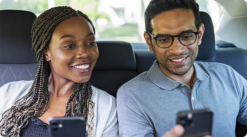 Two people sitting side by side in a car, smiling and looking at their smartphones.
