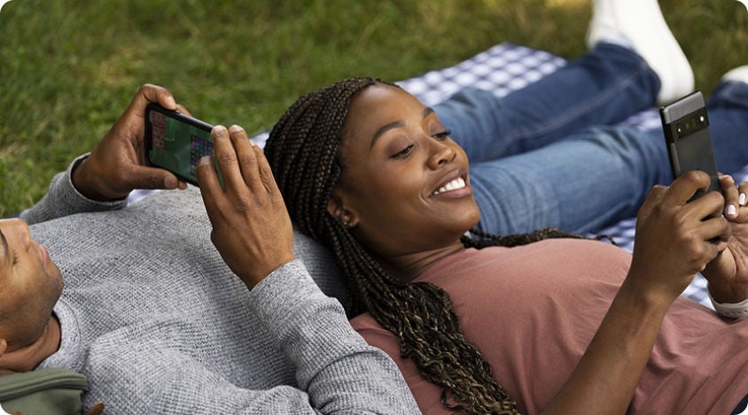 Two people relaxing on a blanket outdoors, each using a smartphone and smiling while playing mobile games.