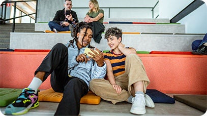 Four people, two are sitting on a big stair in the background and two are sitting in a first row. Both couples are using a phone.