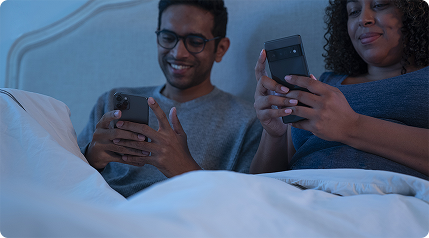 Two people sitting in bed at night, smiling while using their smartphones side by side.