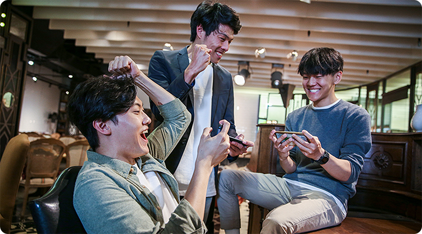 Three people in a lounge-style setting smiling and reacting while playing games on their smartphones.
