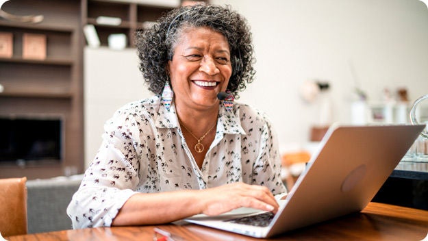 A person with curly hair, wearing a patterned shirt, working on a laptop at a wooden table.