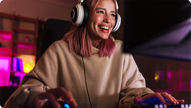 A person wearing large headphones and a beige hoodie, seated at a desk using a computer with colorful lighting in the background.