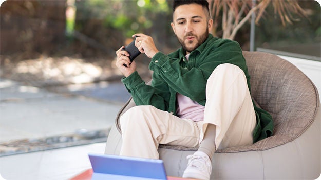 A person sitting outside on a bean bag, holding a game controller, and playing videogames on a tablet.