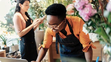 Two people in a flower shop, one in the background looking at a cell phone, one in the front working on a laptop.