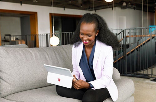A woman in formal clothes sits on a couch, legs crossed, and smiles while using her Microsoft tablet.