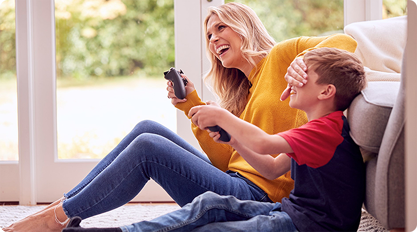 Woman playing a console game with a child who covers his eyes playfully while laughing on the floor at home. 