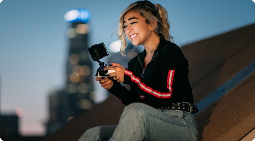 Woman smiling and sitting on a roof playing a mobile game with a city in the background