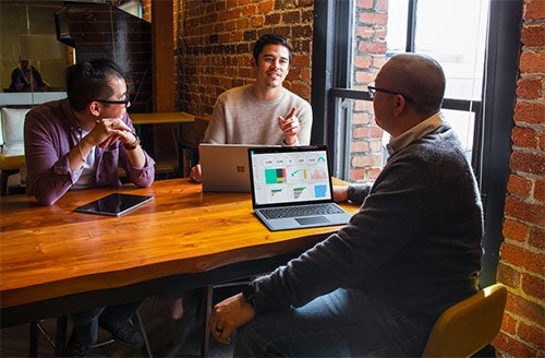 Three men sit at a cafe and converse while working on laptop computers.