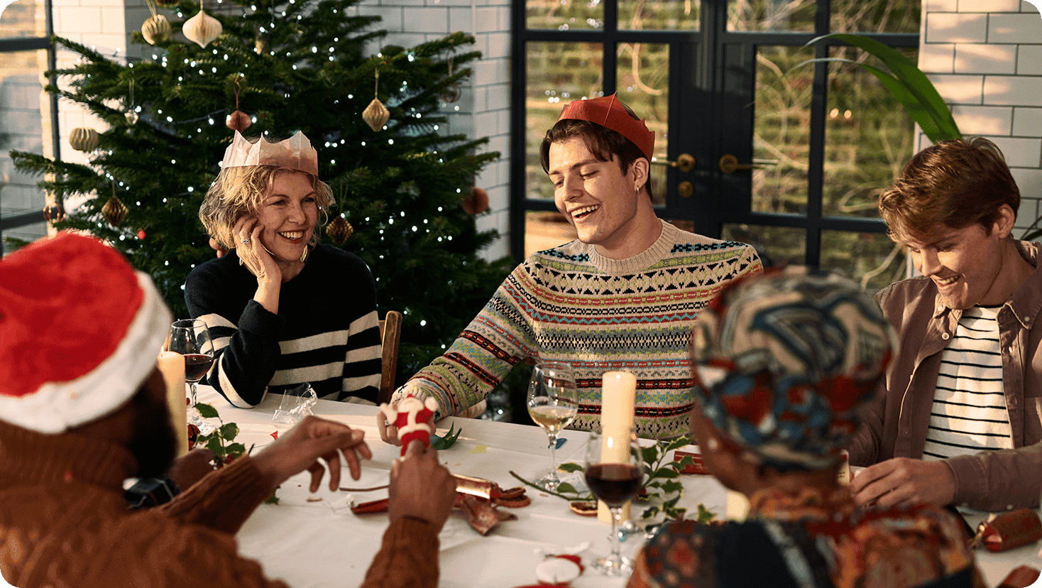 Group celebrating a holiday gathering around a dining table beside a decorated tree and seasonal decorations.