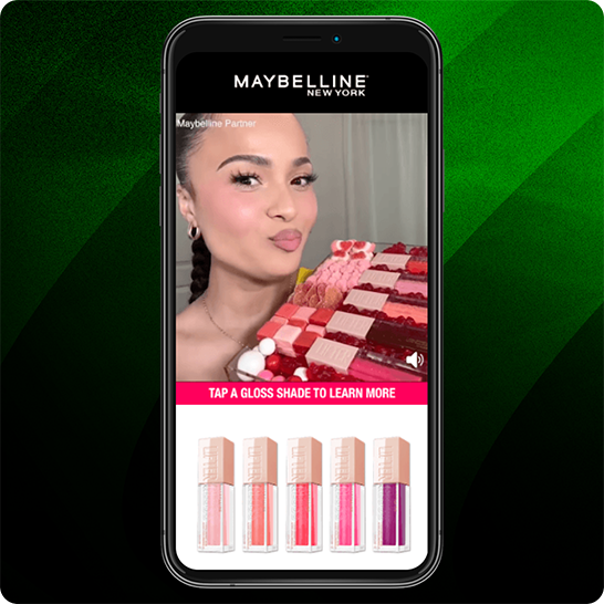 Smartphone displaying a Maybelline New York interactive ad with a pink banner reading “Tap a gloss shade to learn more” and five lip gloss shades shown at the bottom, set against a green striped background.
