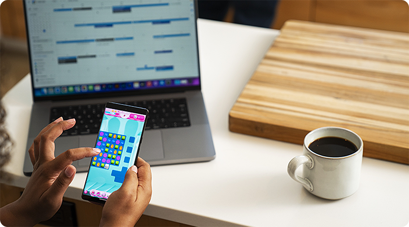 Person holding a smartphone and playing a colorful puzzle game at a desk with a laptop and coffee.