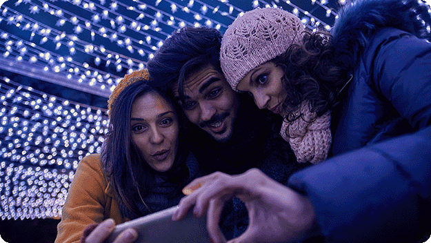 People gathered under illuminated holiday decorations while capturing a photo on a smartphone.