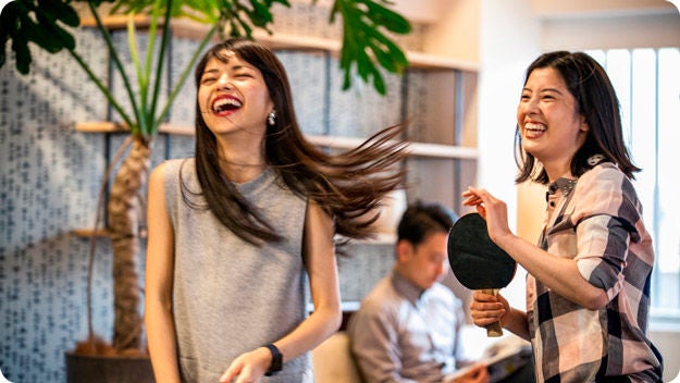 Two people laughing while playing ping pong indoors. One person is holding a paddle. A third person is seated in the background.