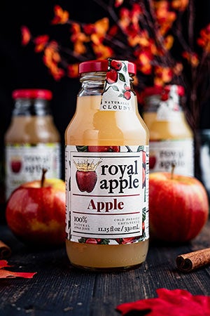 Three bottles of Royal Apple Juice stand in a fall setting composed of apples, a tree, and cinnamon sticks.