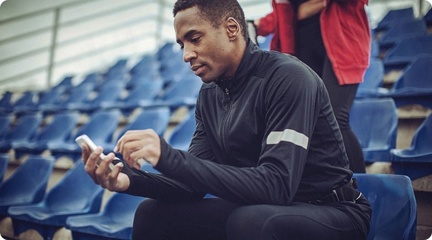 Person seated in an outdoor stadium, looking at their smartphone while dressed in athletic wear.