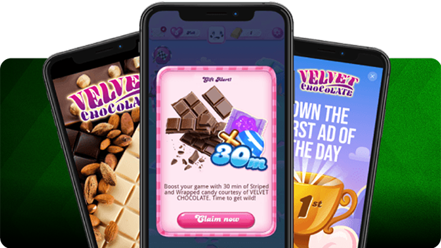 Three smartphones displayed against a green background: the center phone shows a Candy Crush gift alert with chocolate bars and a “30m” boost icon, while the side phones feature Velvet Chocolate ads—one with assorted chocolate pieces and almonds, the other with a golden trophy and text promoting an ad of the day. 