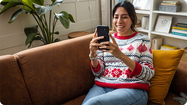 A person sitting on a couch indoors, looking at a cellphone and smiling.