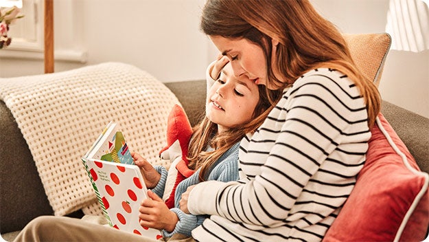 An adult and a child are sitting on a couch. The adult is wearing a striped shirt and the child is holding an open book with a colorful cover featuring red polka dots.