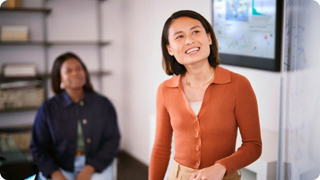 Two peoplein a modern office, one seated in the background and one standing, with digital screens displaying data.