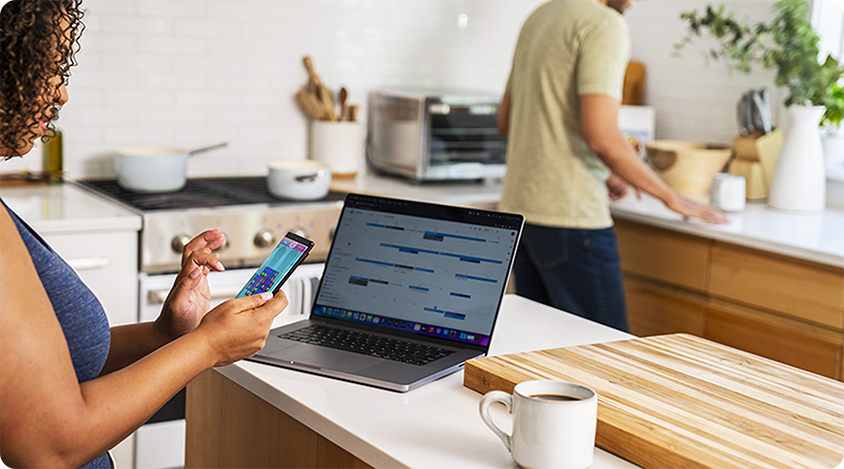 Person standing in a kitchen playing Candy Crush Saga on their smartphone near an open laptop and a cup of coffee.