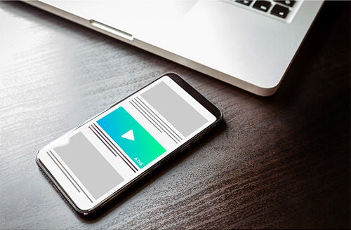 A cellphone rests on a wooden table next to a partial view of a laptop. The screen shows a mockup of an ad placement and site layout using color blocks.