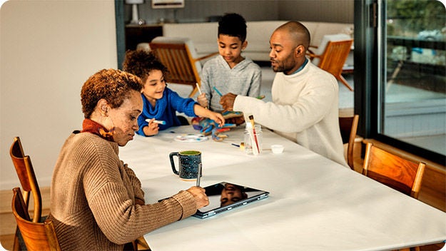 A family of four is seated around a dining table. A person with short curly hair is sitting at the end of the table, looking at a tablet. Two children are engaged in an art activity with colored markers and paper, while an adult assists them.