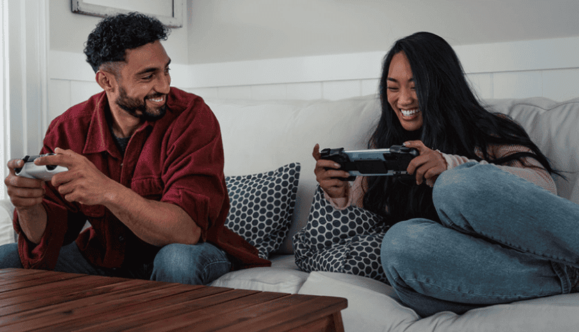 Pair of friends laughing and gaming together on a couch, one with a controller and another with a handheld gaming device