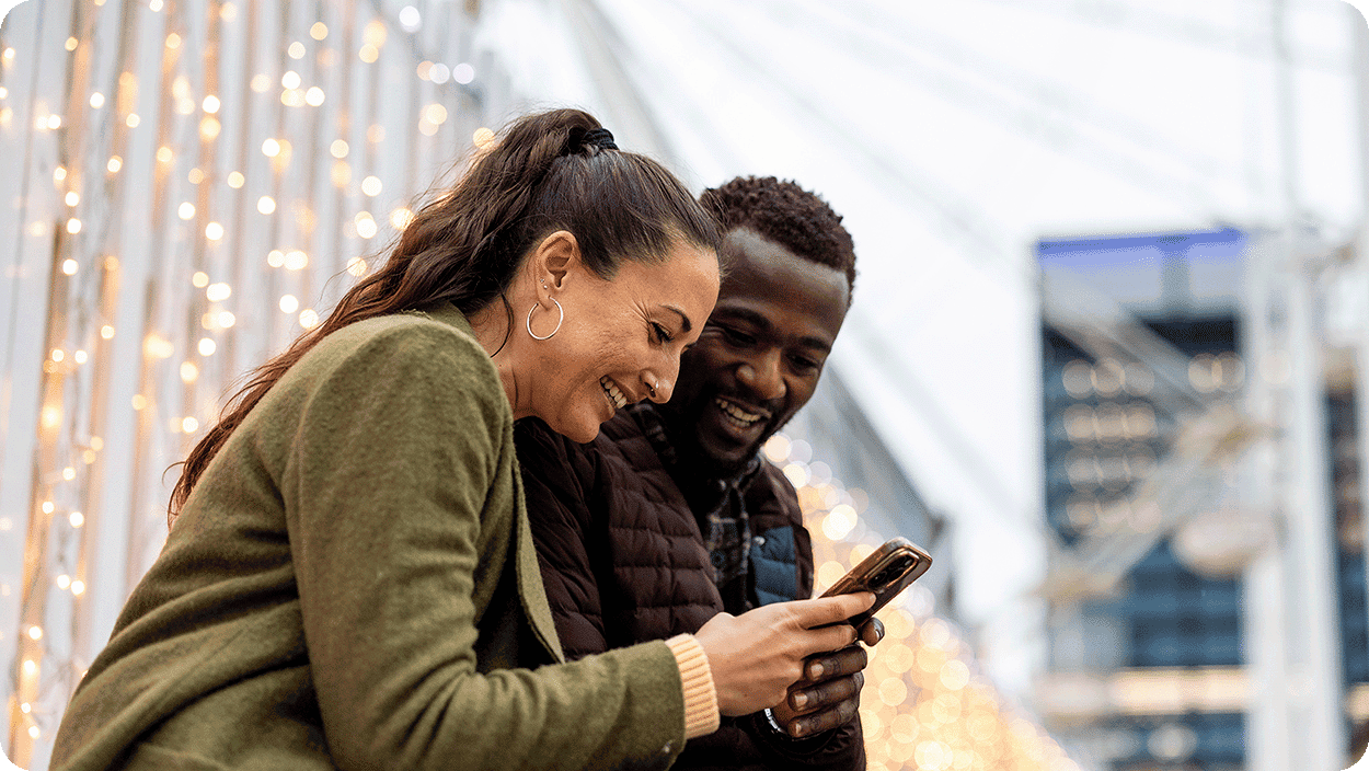Two people standing outdoors and viewing a smartphone together. Warm string lights and an urban setting are visible.