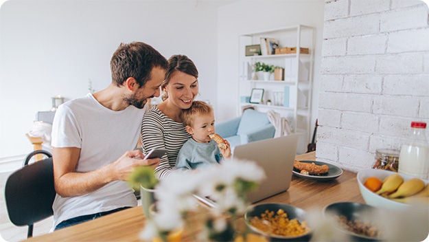 A family of three is sitting at a wooden dining table in a modern, bright kitchen. The father is holding a smartphone, while the mother is holding their child on her lap and looking at a laptop screen.