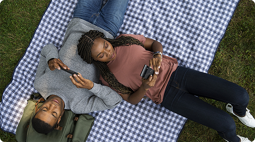 Two people lying on a blanket outdoors, each using a smartphone while relaxing side by side.