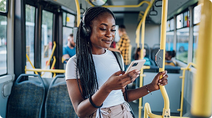 Woman standing on a bus holding a pole and smiling at her smartphone while wearing headphones.