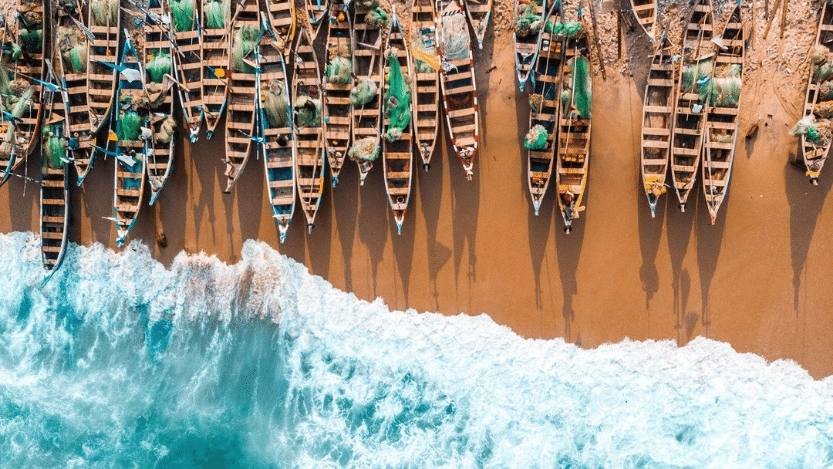 An aerial view of fishing boats on the Cape Coast of Ghana moored on sandy shore while the aqua colored tide breaks near them.