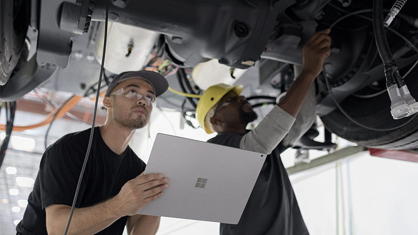 Two mechanics stand under a car fixing it while holding a Microsoft tablet.