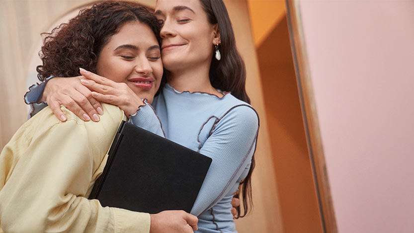 Two women embrace with closed eyes and one of them holds a laptop computer.