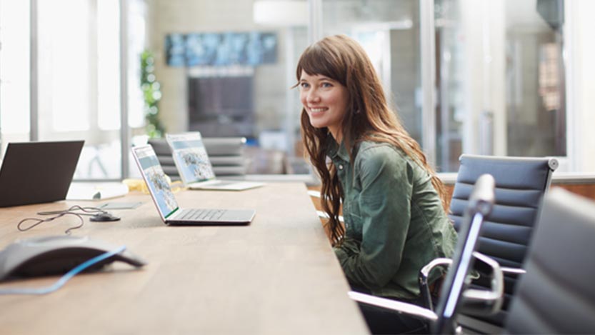 A woman sits at a desk in front of a laptop and smiles away from the camera.