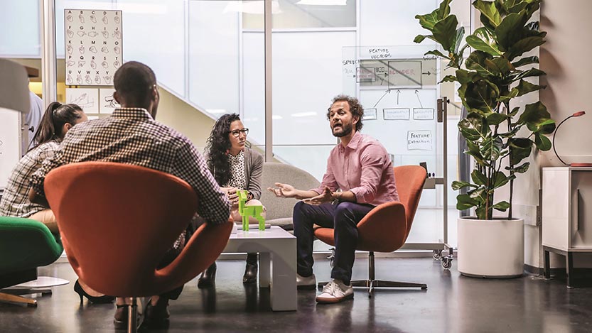 A group of people sitting in an office around a coffee table discuss a topic.