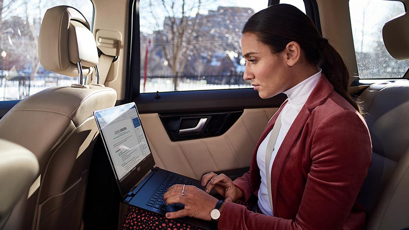 A woman sitting in the back of a car works on her laptop computer