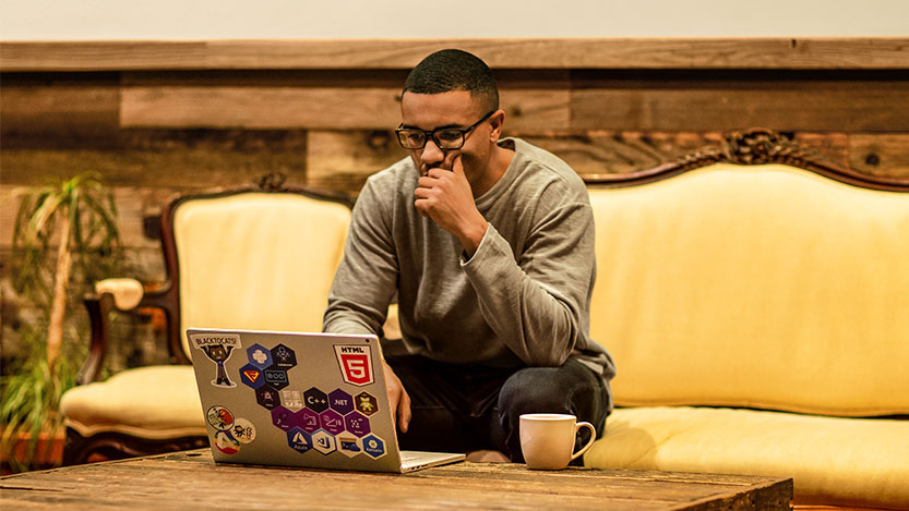 Man sitting down on a couch and working on his personal computer while drinking coffee.