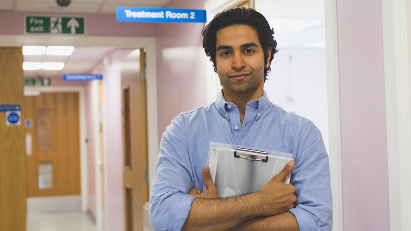 A man in a hospital setting holds a chart and smiles while looking at the camera.