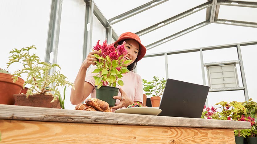 A woman inside a greenhouse is holding a plant while looking at a laptop placed on a desk.